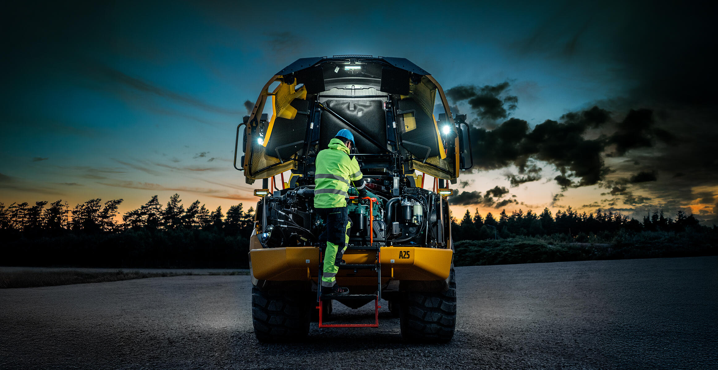 A Volvo technician is working inside the engine compartment of a Volvo Articulated Hauler at night in a quarry environment. Lights around the machine light up the area the engineer is working in.