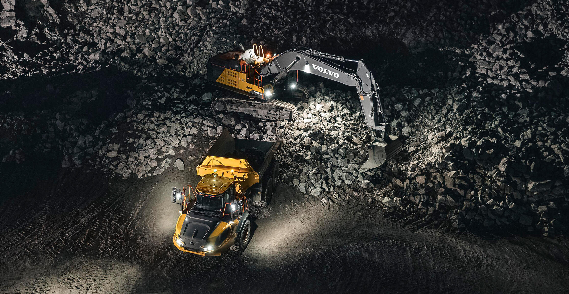A Volvo Articulated Hauler is working at night in a quarry being loaded by a Volvo excavator. Work lights on the machines light up the surrounding area.