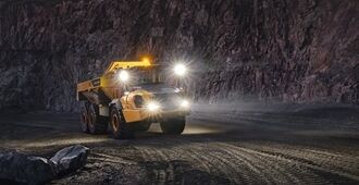 En rammestyret Volvo A50 dumper med fuld affjedring kører op ad en transportvej i et stenbrudsmiljø om natten med lygterne tændt.