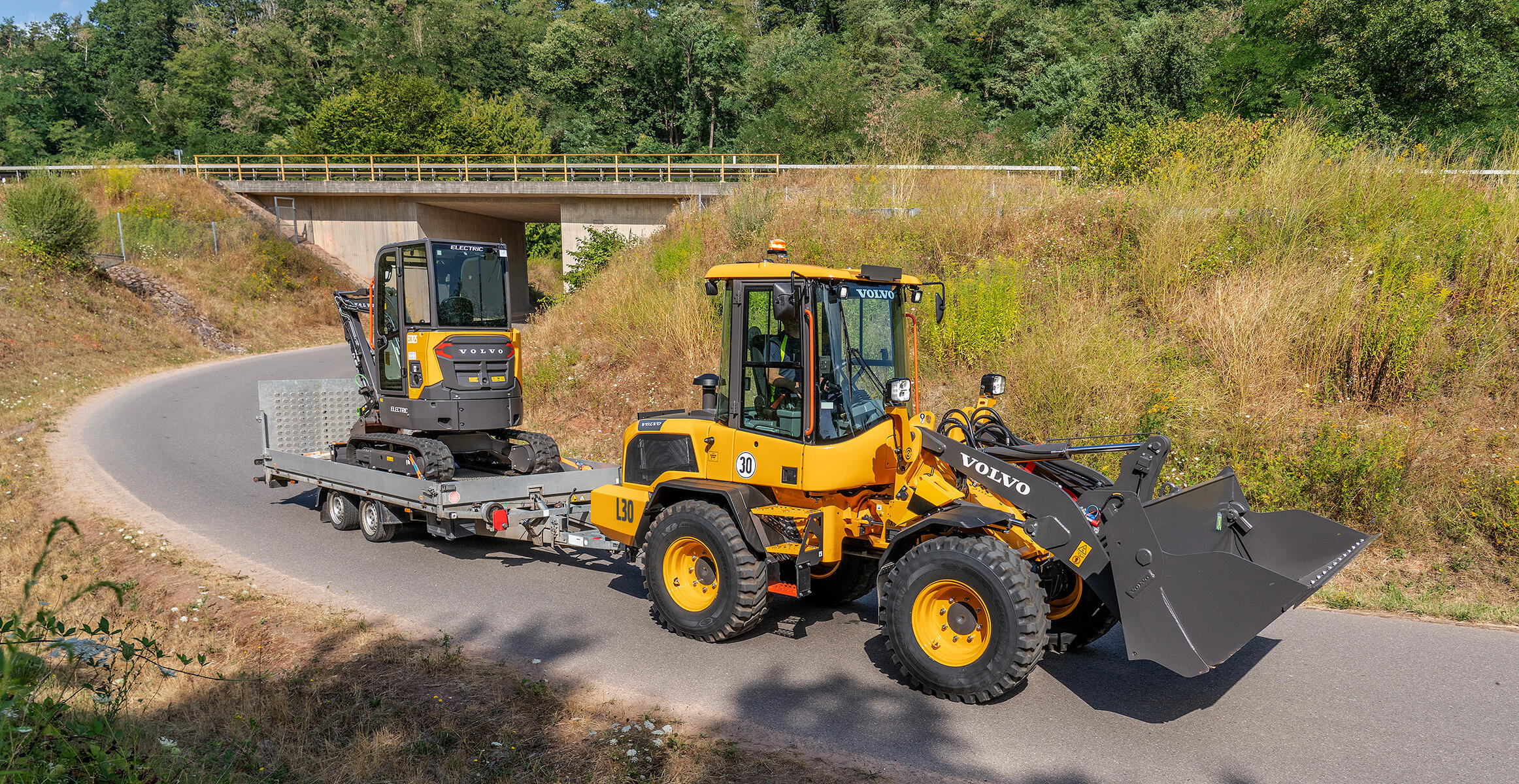 Volvo L30 Compact Wheel Loader towing a trailer carrying a compact excavator.