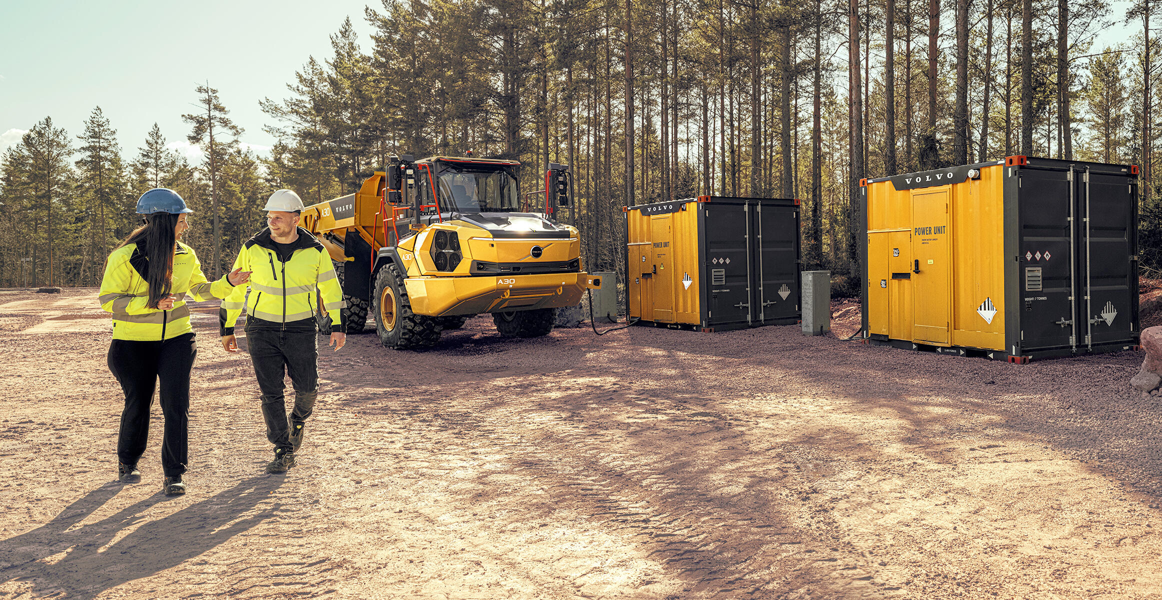 Deux personnes sur un chantier de terrassement marchant devant un nouveau tombereau articulé électrique Volvo A40 Electric et une unité mobile d'alimentation Volvo PU500