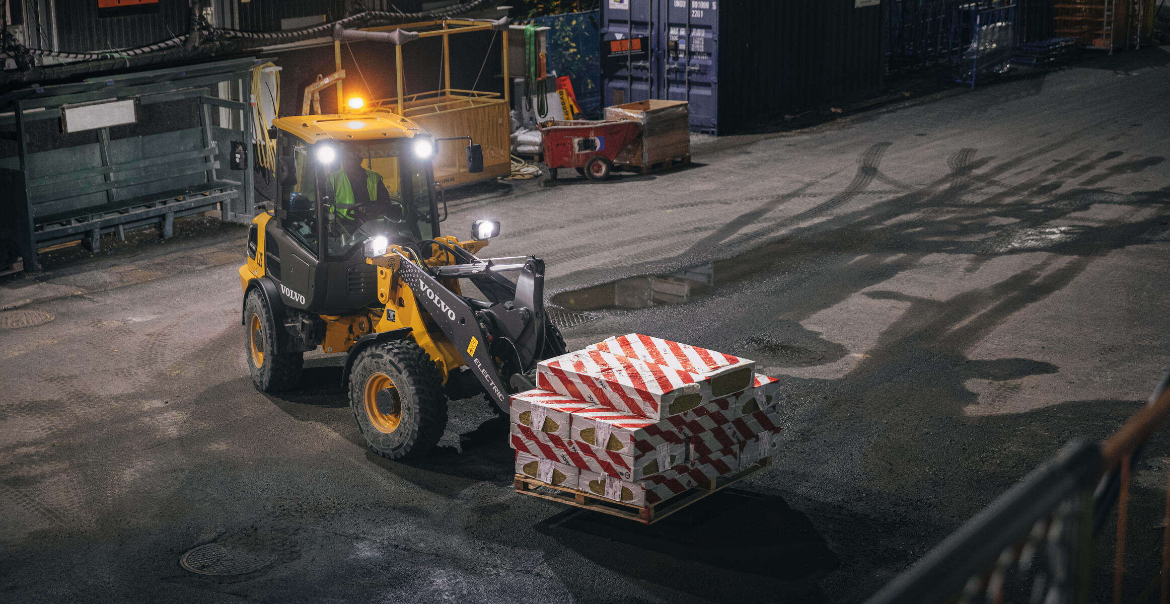 Widok z przodu przedstawiający ładowarkę kompaktową L25 transportującą sprzęt przy użyciu wideł do palet