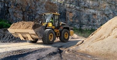 Volvo L120 Electric wheel loader dumping grayish-brown aggregate from raised bucket into a dump truck bed on an outdoor urban construction site