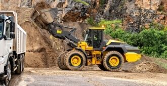 A Volvo L120 Electric Wheel Loader operates in an industrial setting, moving materials with its front-mounted bucket filling a bed truck in quarry site.