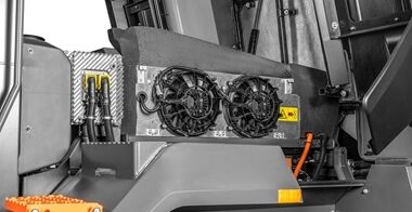 Close-up of the Volvo L120 Electric Wheel Loader's cooling system compartment, featuring dual fans, high voltage warning label reading "DANGER HIGH VOLTAGE," yellow connectors, black hoses, and an orange cable, highlighting its low-maintenance electric design.