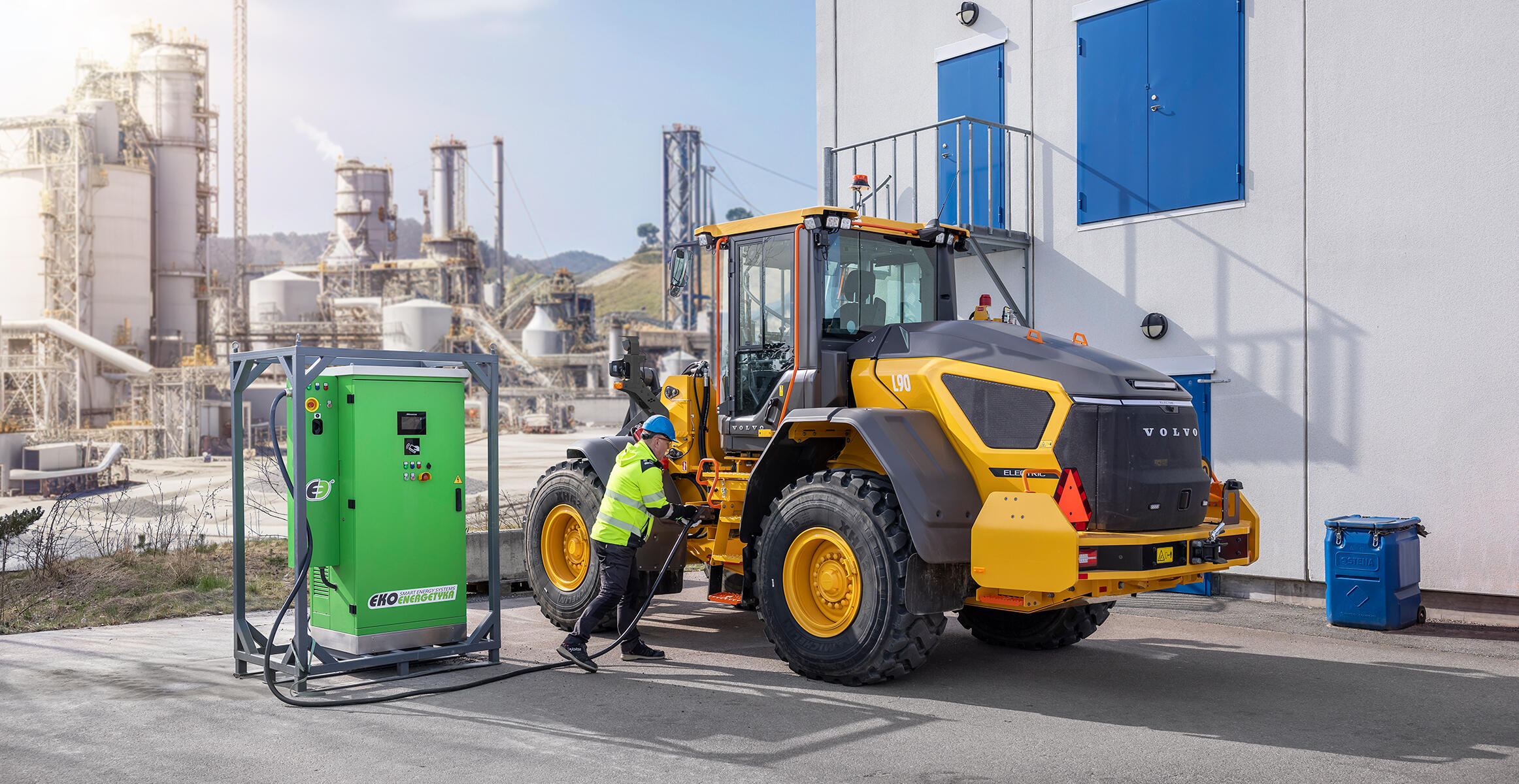 Operator podłącza elektryczną ładowarkę kołową L90 Electric do stanowiska w celu naładowania akumulatora.