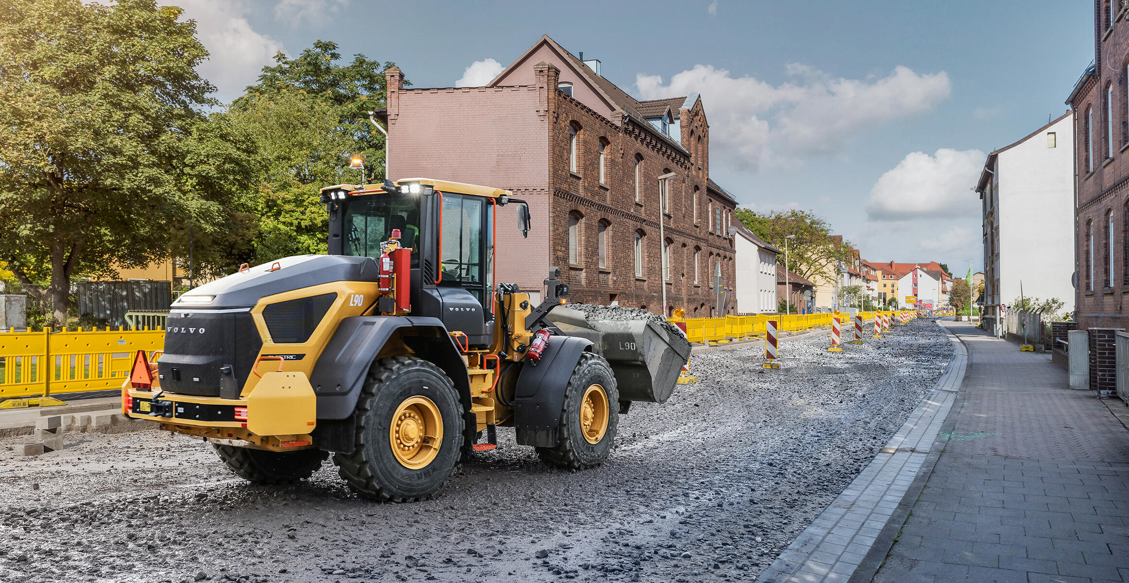Volvo L90 Electric læssemaskine med fuld grusskovl, som manøvrerer gennem et boligbyggeri under en klar, blå himmel.