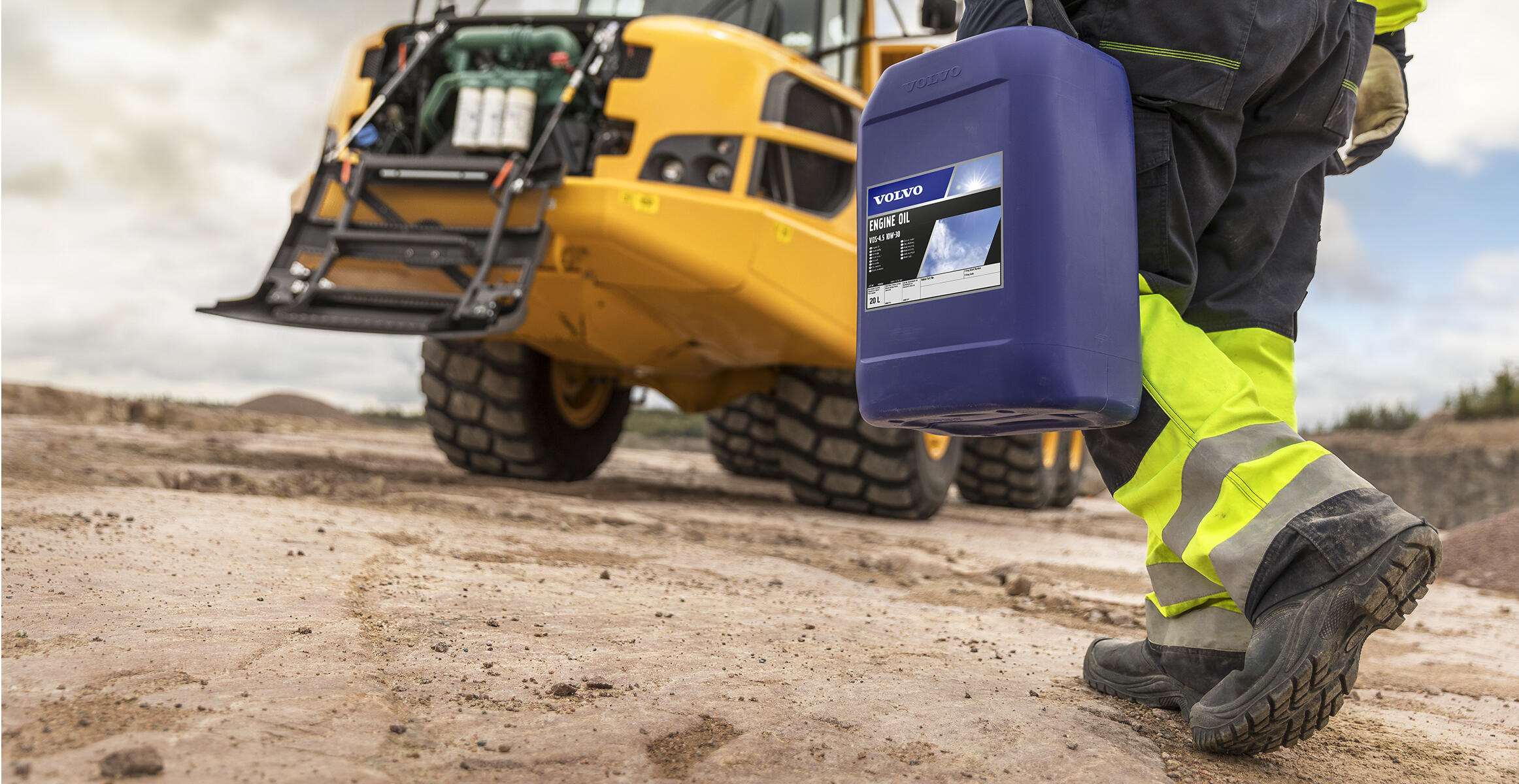 Worker carrying Volvo engine oil container at construction site with yellow excavator in background