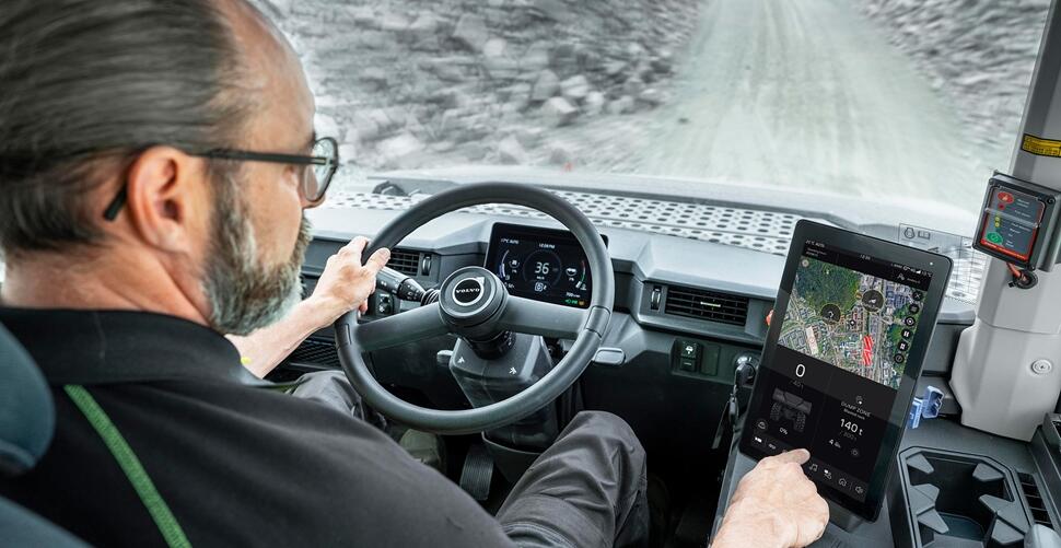 Operator driving a Volvo articulated dump truck on a gravel haul road, viewed from inside the cab.