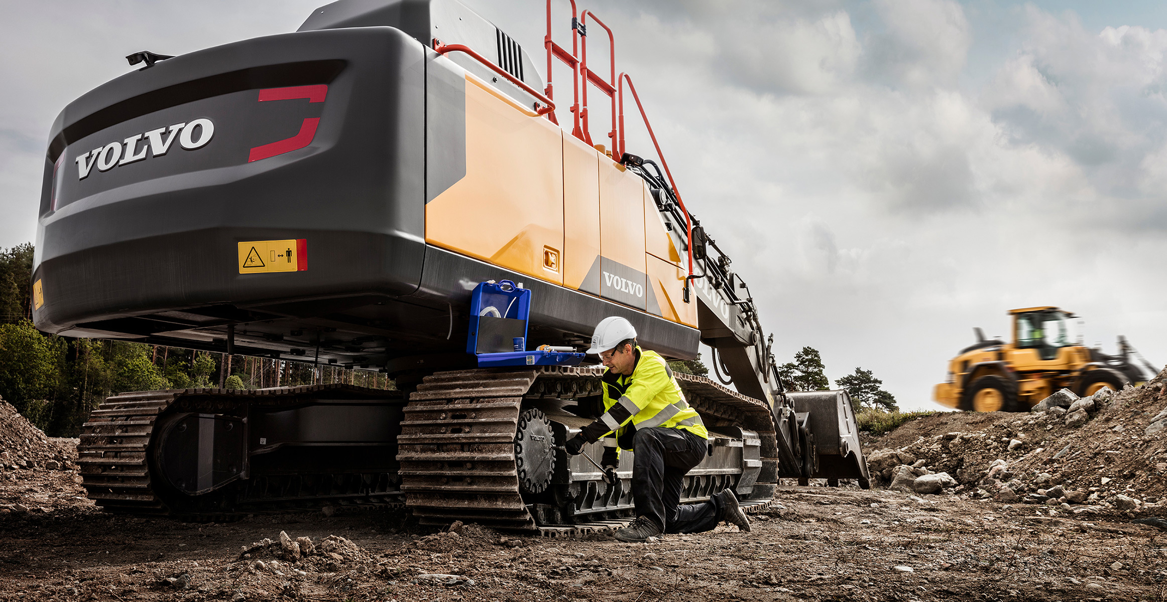 A service tech providing maintenance on a Volvo crawler excavator with a wheel loader working in the background.
