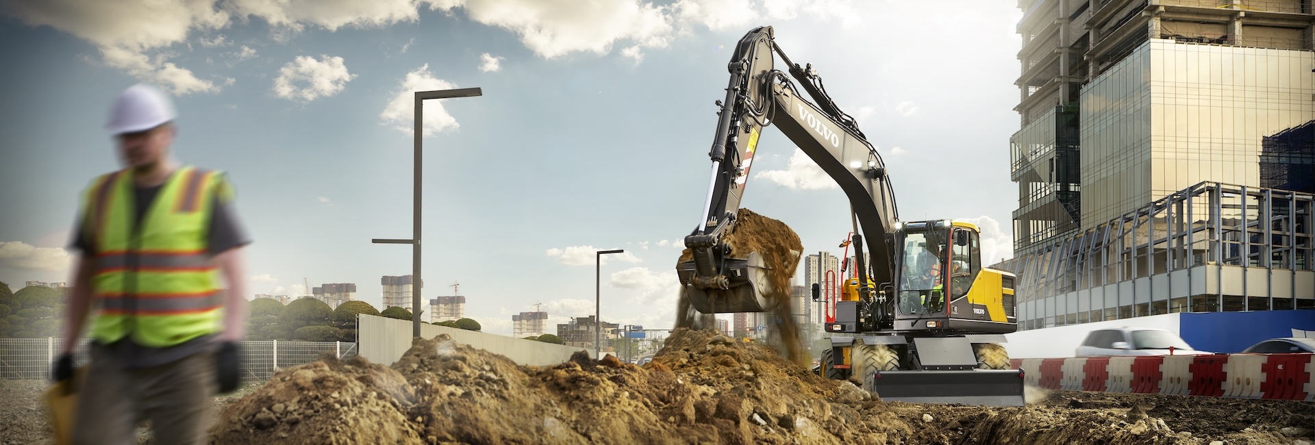 Volvo wheeled excavator working in the summer heat in the background with a construction worker walking nearby on the left.