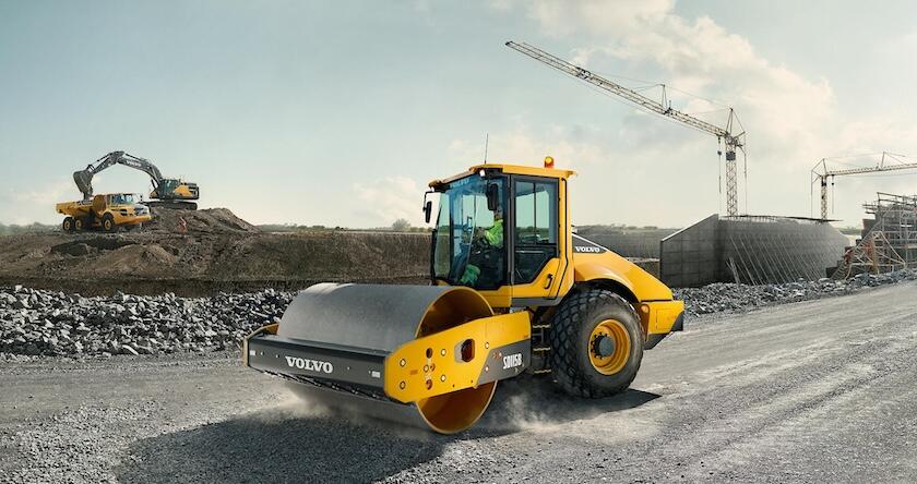 A Volvo SD115B soil compactor compacting a gravel road with a Volvo excavator and ADT working together in the background.