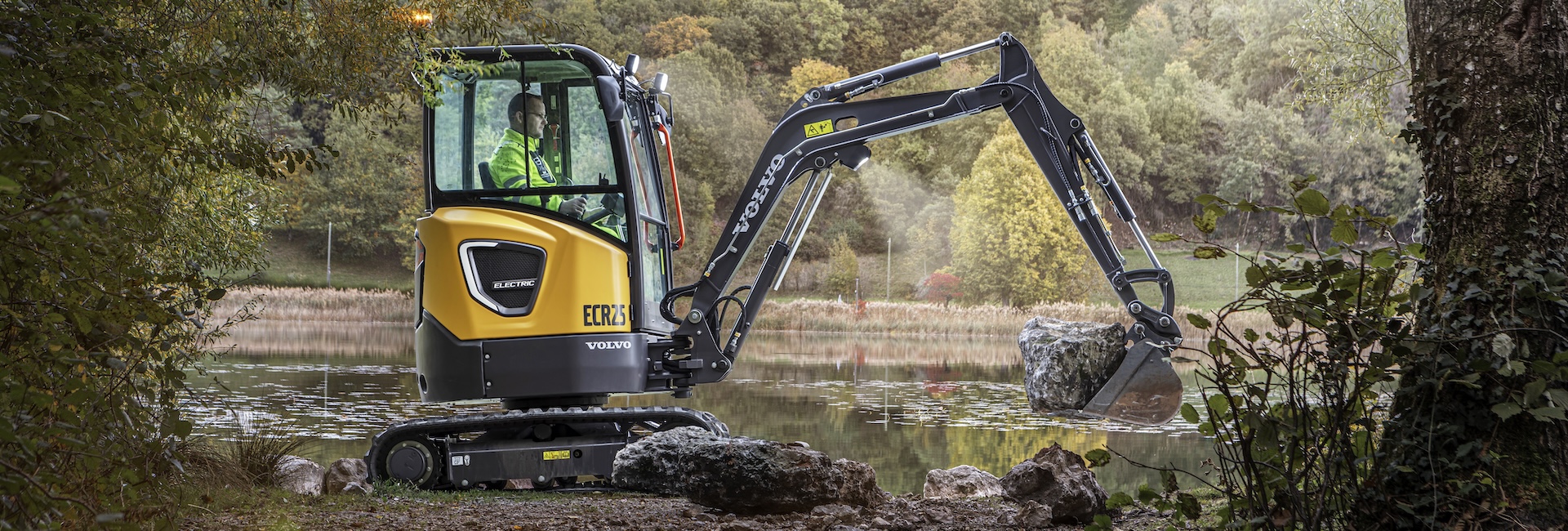A Volvo ECR25 Electric excavator landscaping by moving a boulder next to a pond.