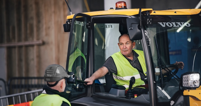 A Volvo electric equipment operator in the cab talking to a crew member next to him.