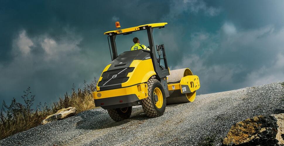 A Volvo SD75B soil compactor operating up an incline with a rain storm in the distance.
