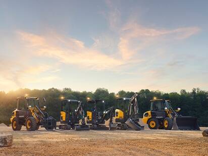 The lineup of Volvo electric compact wheel loaders and electric compact excavators on dirt with their lights on.