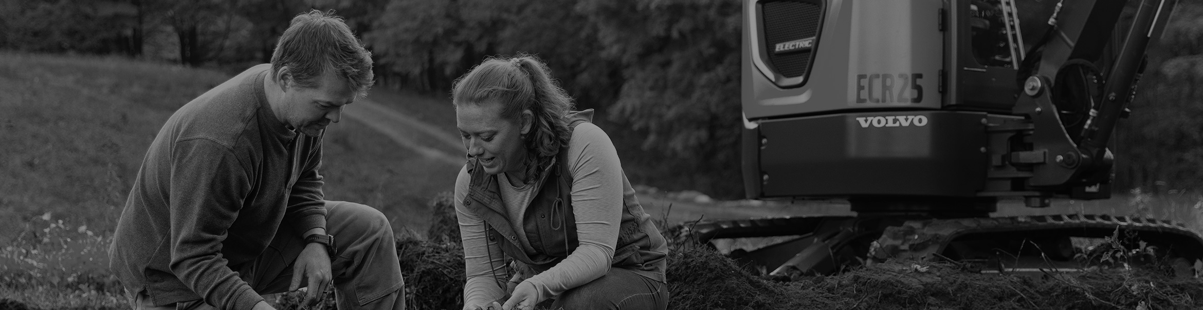A man and a woman who own a farm crouch in front of their Volvo electric excavator.