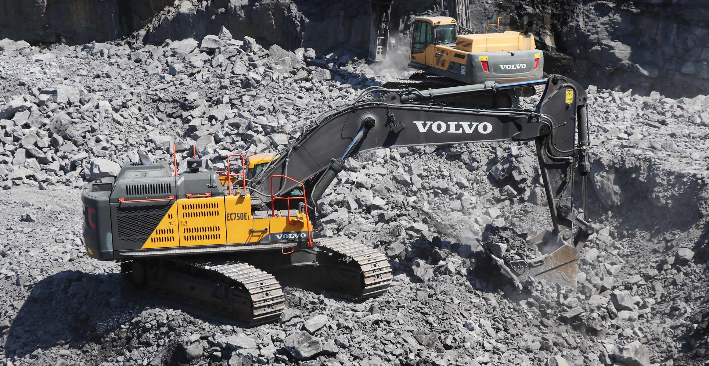 A Volvo excavator breaking rock in the background with a Volvo EC750E excavator moving the rock in a quarry.