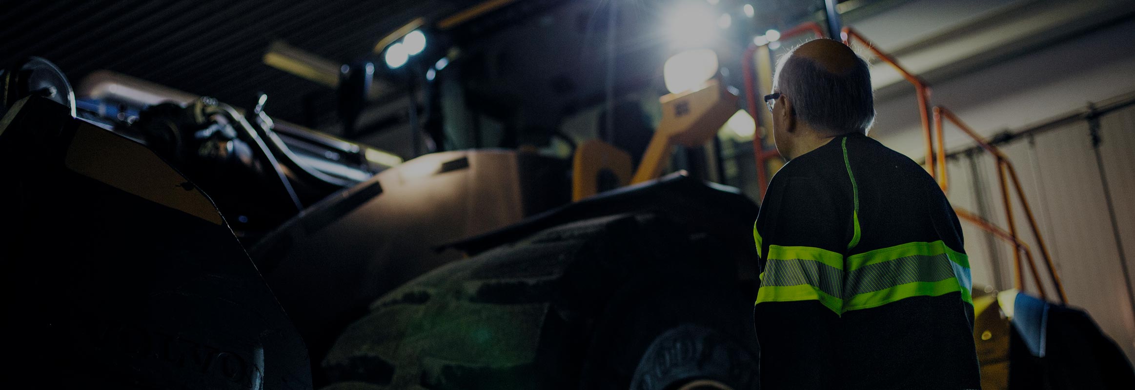 A man looking up at a Volvo wheel loader with its lights on in a darkened room.