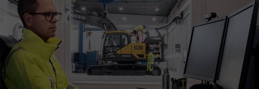 Technicians servicing a Volvo EC230 excavator in the background, with a tech in the foreground at a computer.