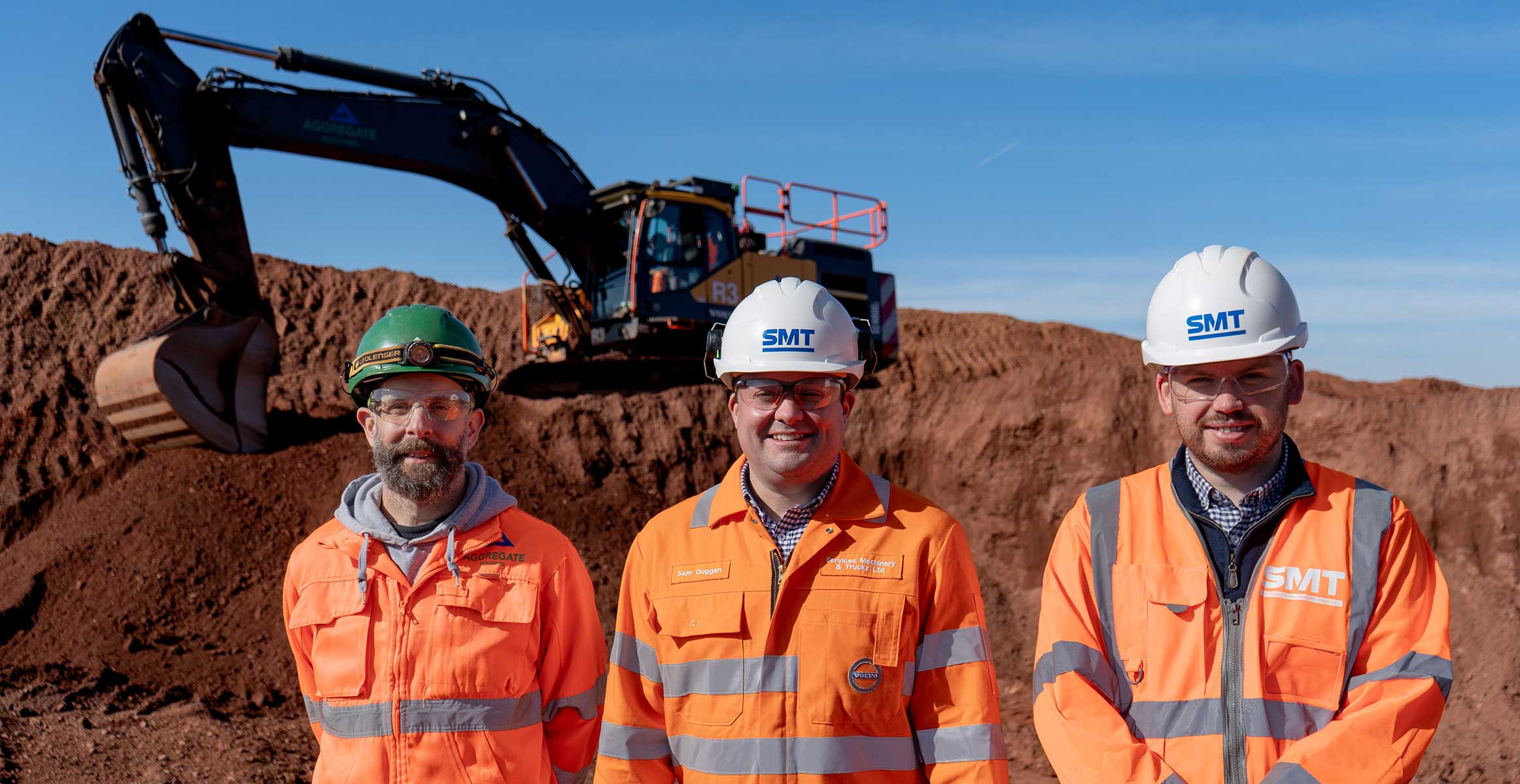 Trois ouvriers en tenues haute-visibilité et casques SMT posant devant une excavatrice sur chantier